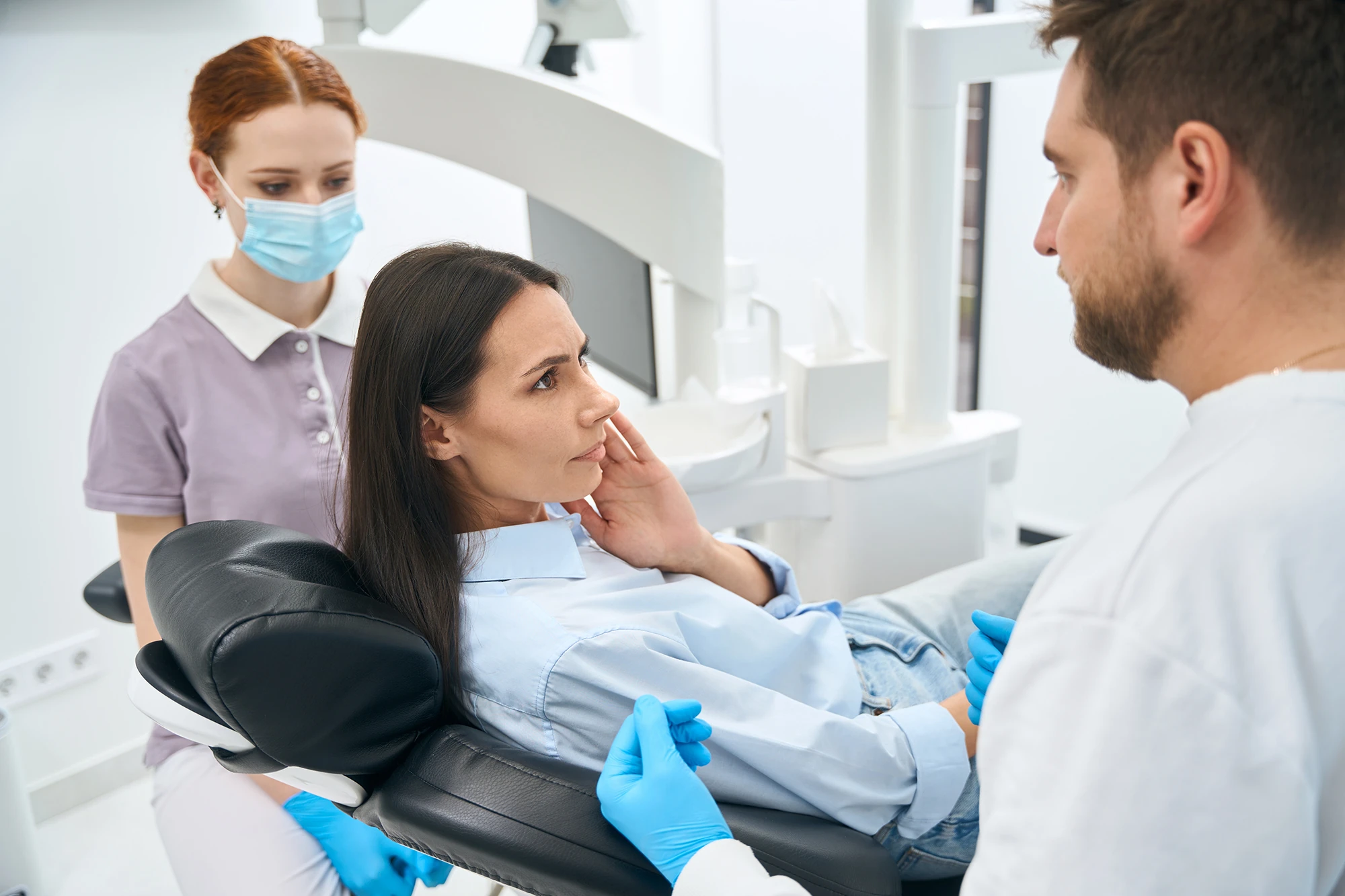 Patientin im Zahnarztstuhl, Zahnarzt berät, Assistenz mit Maske daneben